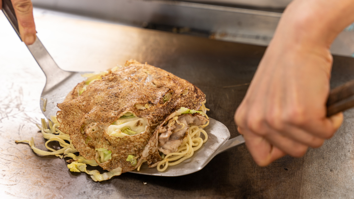 ライブキッチンの鉄板で広島風お好み焼きを、コテでひっくり返そうとしている様子