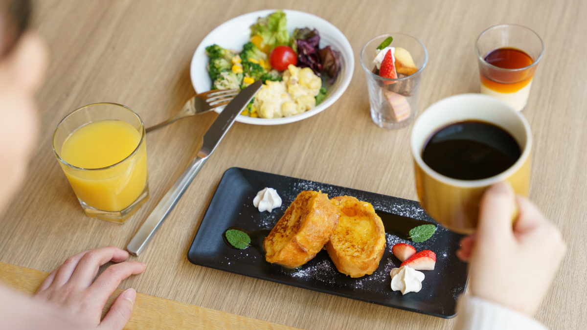 朝食を食べる人の手元とフレンチトースト、サラダ、オレンジジュース、コーヒーの写真