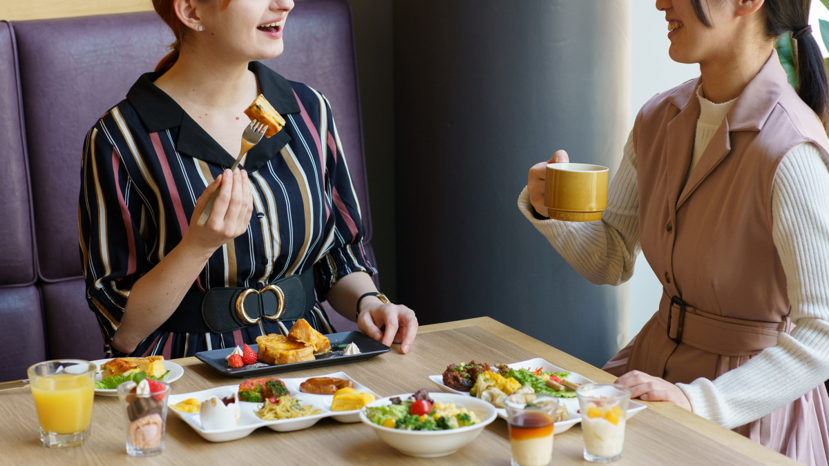 2人の女性が朝食ビュッフェを楽しんでいる写真、一人がキッシュをフォークで持ち、もう一人がカップを持っている